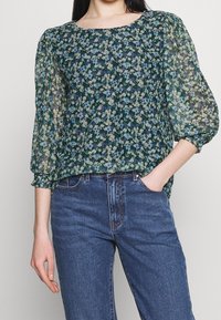 Hedvábná blůza s květinovým vzorem v tmavě zelené barvě, s průsvitnými nabíranými rukávy a čtvercovým výstřihem. V kombinaci s modrými džíny, které zdůrazňují uvolněný střih.