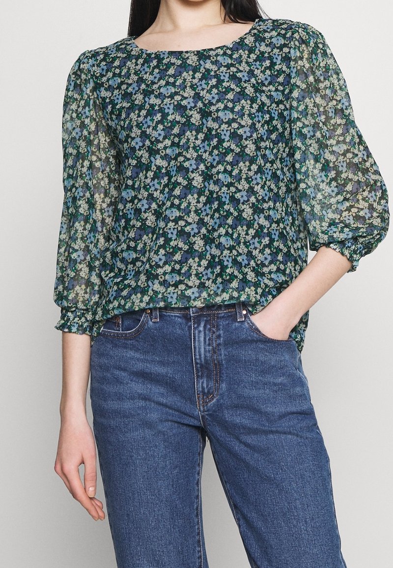 Hedvábná blůza s květinovým vzorem v tmavě zelené barvě, s průsvitnými nabíranými rukávy a čtvercovým výstřihem. V kombinaci s modrými džíny, které zdůrazňují uvolněný střih.