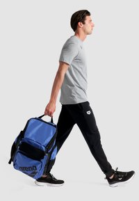 Mochila deportiva azul y negra con un bolsillo de malla y un gran logo, llevada por una persona que viste una camisa gris y pantalones negros, caminando.