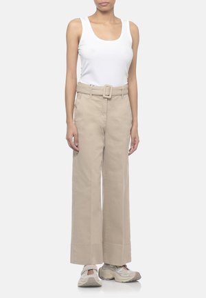 Donna con top bianco senza maniche, pantaloni beige a gamba larga e vita alta con cintura e sneakers beige, in piedi davanti a uno sfondo semplice.