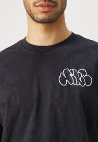 Camisa de manga larga de algodón negro con un logo bordado en blanco en el pecho. Textura suave, con un diseño de cuello redondo y un corte relajado.