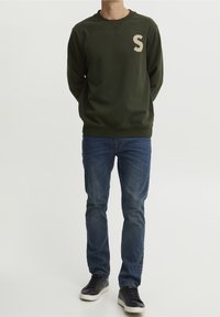 Zielona bluza z beżowym logo 'S', z okrągłym dekoltem, ściągaczami na rękawach, w zestawie z niebieskimi dżinsami i czarnymi sneakerami.