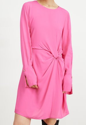 Frau trägt ein langärmeliges rosa Kleid mit einem gedrehten Knotendetail an der Taille und langen Manschetten mit Knöpfen.