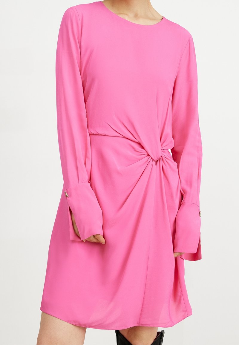 Femme portant une robe rose à manches longues avec un détail de nœud torsadé à la taille et de longues poignets avec boutons.
