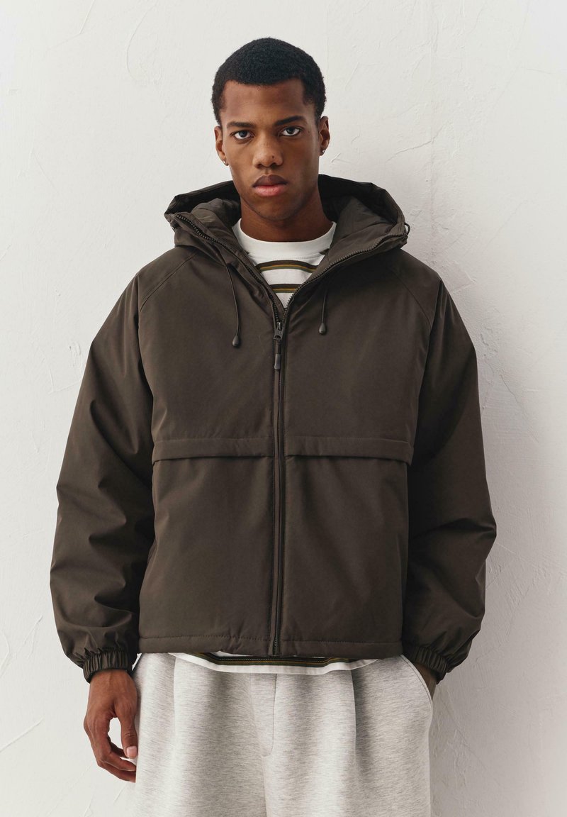 Bruine capuchonjas met een ritssluiting aan de voorkant, elastische manchetten en een horizontale borstzak, gedragen over een wit shirt met strepen.