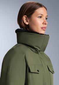 Olivno zelena vodoodporna jakna s visokim ovratnikom in črnim umetnim krznom, dvema žepoma na prsih ter oznako RECCO na rokavu.