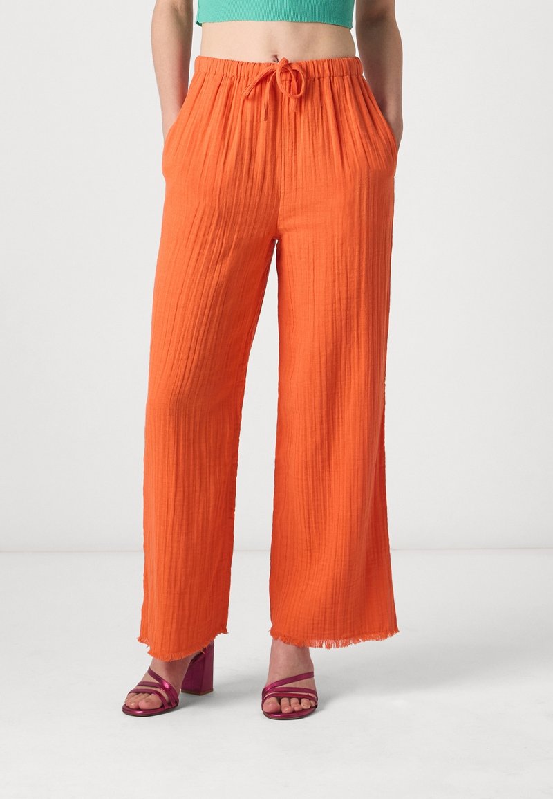 Pantalons orange à jambes larges avec un tissu texturé et un ourlet effiloché, dotés d'une taille élastique avec un cordon de serrage. Associés à des sandales roses à lanières.