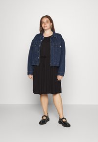 Vestido preto com um cinto na cintura, sobreposto por uma jaqueta de ganga azul escura com botões e bolsos laterais. Sapatos pretos com um salto baixo.