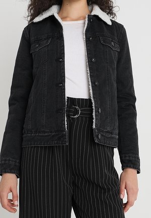 Kvinde iført en sort denimjakke med hvid fleecekrave, hvid ribbet top og sorte højtaljede bukser med hvide striber og bælte.