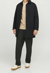 Tmavě námořnický kabát s límcem, zapínáním na knoflíky a dlouhými rukávy, nošený přes béžový pletený svetr a černé kalhoty, doplněný bílými teniskami.