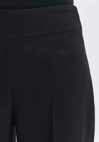Gros plan d'un pantalon noir en tissu texturé à taille haute montrant la ceinture et les détails de la couture latérale.