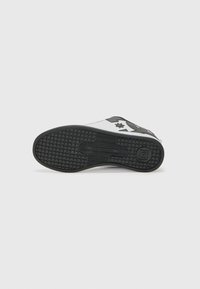 Zapatilla con suela de goma negra, parte superior blanca y acentos en negro contrastantes. Presenta una superficie texturizada y un detalle del logo de una estrella.