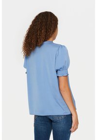 Blusa blu a maniche corte con maniche a sbuffo e polsini arricciati, caratterizzata da una vestibilità morbida e una texture liscia. Indossata con jeans blu.