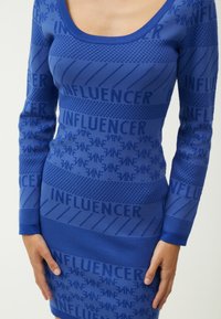 Blaues Strickkleid mit Rundhalsausschnitt, langen Ärmeln und einem sich wiederholenden "INFLUENCER"-Muster mit unterschiedlichen Texturen.