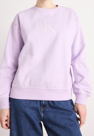 Henkilö, jolla on yllään vaaleanvioletti Calvin Kleinin collegepaita ja siniset farkut, katsoo eteenpäin kädet rentoina sivuilla.