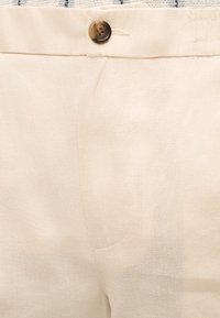 Κοντινό πλάνο μπεζ λινών παντελονιών με καφέ κουμπί και ορατές ραφές κοντά στη μέση και την περιοχή του φερμουάρ.