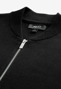 Černá svetr s zipem až ke krku, s žebrovaným límcem, který má stříbrný zip a tkaninovou texturu. Na etiketě je uvedeno "NEXT" s přiloženými údaji o údržbě.