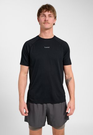 Man met snor die een zwart sportshirt met korte mouwen en grijze gelaagde shorts draagt, staand tegen een effen lichte achtergrond.