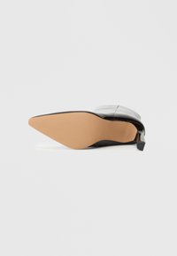 Sapato de salto alto preto com bico fino, com um cabedal em couro liso de verniz, sola de borracha bege e um design de salto esguio e angular.