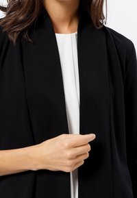 Schwarze, offen vorne stehende Jacke aus einem glatten, leichten Stoff. Das kragenlose Design verfügt über drapierte Schultern und einen entspannten Schnitt.