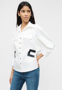 Γυναίκα που φοράει λευκό πουκάμισο με μαύρες λεπτομέρειες στις πλευρές και μπλε τζιν, στέκεται με το ένα χέρι στη μέση.