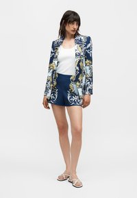 Γυναίκα που φοράει ένα μπλε και λευκό σακάκι με μοτίβο και ταιριαστά σορτς πάνω από λευκή κοντομάνικη μπλούζα, στέκεται με σανδάλια σε λευκό φόντο.