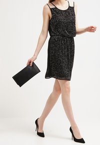 Czarna sukienka bez rękawów z srebrnymi pionowymi liniami, elastycznym pasem, przezroczystą narzutą i pasującą czarną kopertówką. Modelka w czarnych szpilkach.