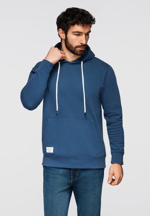 Homme portant un sweat à capuche bleu avec des cordons blancs et un jean bleu, ajustant le col de sa capuche, tourné légèrement vers la gauche sur un fond uni.