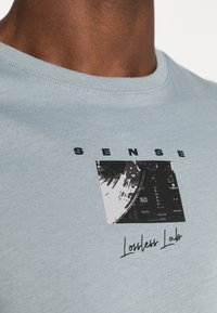 T-shirt en coton bleu clair avec un col rond, présentant un design graphique noir qui comprend du texte et une image abstraite.