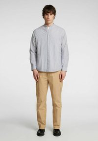 Juostuota šviesiai mėlyna ir balta marškinėlių chemise, su mandarinų apykakle, ilgomis rankovėmis ir laisvu siluetu, derinama su smėlio spalvos kelnėmis.