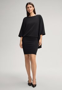 Robe noire avec un haut ample à manches chauve-souris et une jupe ajustée. Tissu lisse de couleur unie, portée avec des escarpins noirs.