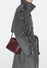 Person trägt einen grauen Wollmantel mit hohem Kragen und schwarzen Knöpfen, hält eine burgunderrote geflochtene Leder-Umhängetasche, Hand in der Tasche.