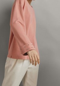 Person wearing a long-sleeve pink top with metal studs on cuffs and light beige pants against a plain gray background.