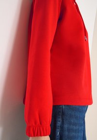 Czerwona bluza z kapturem, ściągaczami na rękawach i luźnym krojem. Połączona z niebieskim denimem, ukazująca gładką fakturę tkaniny.