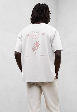 Mężczyzna o ciemnych dredach, ubrany w białą koszulkę z różowym nadrukiem palm i napisem "Bo nie ma Planety B", stojący na jednolitym tle.