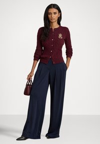 Bordo krāsas raustītas adījuma kardigans ar zelta logotipu, kopā ar plašām tumši zila krāsas biksēm. Modelis tur rokās mazu bordo krāsas rokassomiņu, valkā melnus apavus.