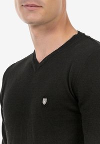 Schwarzer V-Ausschnitt-Pullover aus strukturierter Stoffe, mit einem kleinen, schildförmigen Logo auf der Brust, das sein elegantes Design unterstreicht.
