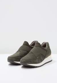 Baskets en daim vert olive avec des sangles élastiques, bout arrondi et semelle blanche. Présentent un léger talon et des détails texturés sur les côtés.