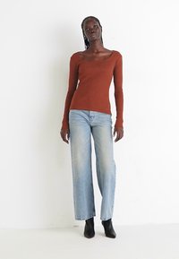 Rode longsleeve top met een ronde hals, gecombineerd met lichtblauwe wijde jeans en zwarte enkellaarzen op een neutrale achtergrond.