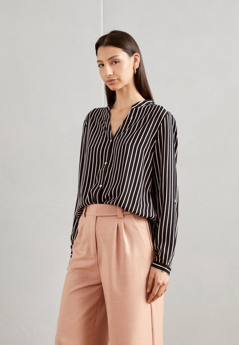 Black and white striped button-up shirt with a v-neck; paired with light pink high-waisted trousers. Smooth fabric, relaxed fit.