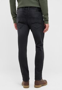 Mężczyzna w ciemnoszarych dopasowanych dżinsach, brązowych skórzanych butach i zielonej bluzce z długim rękawem, stojący na jednolitym jasnym tle.