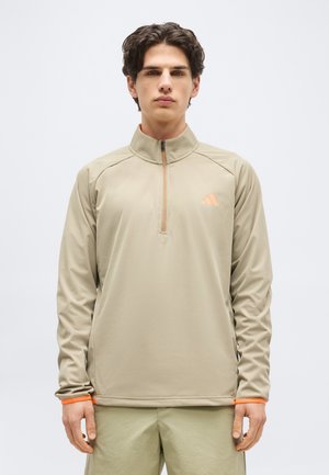 Noormees, kes vaatab otse kaamerasse, kandes beeži värvi neljandiku lukuga džemprit, millel on oranž logo ja ääris, seisab ühevärvilise heleda tausta ees.