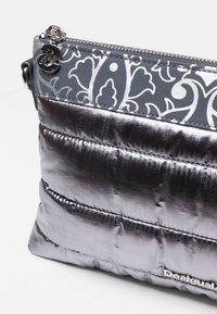 Metaliczna srebrna torebka z pikowaną fakturą. Górna część wykonana z ciemnoszarej tkaniny z wzorem. Zapięcie na suwak i logo marki po boku.