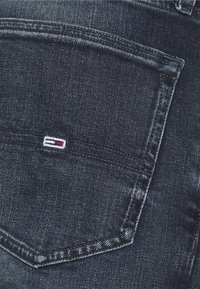 Vaqueros de mezclilla azul oscuro con un bolsillo trasero que presenta un pequeño logo bordado en colores rojo, blanco y azul. Textura desgastada cerca del bolsillo.