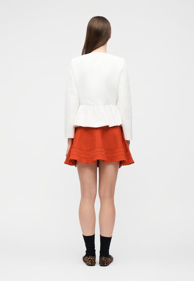 Γυναίκα με μακριά καστανά μαλλιά που φοράει λευκό σακάκι πεπλούμ, πορτοκαλί πλισέ φούστα, μαύρες κάλτσες και παπούτσια μοτίβο λεοπάρ, στέκεται με την πλάτη προς τα εμάς.