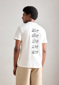 Witte katoenen t-shirt met grafische tekst op de achterkant in verschillende talen, met een ronde halslijn en korte mouwen.