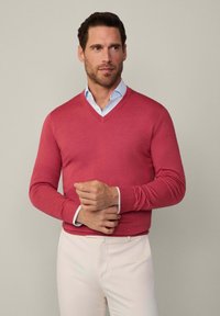 Korallfarbener V-Ausschnitt-Pullover aus weichem Stoff mit langen Ärmeln; gestylt über einem hellblauen Hemd und kombiniert mit cremefarbener Hose.