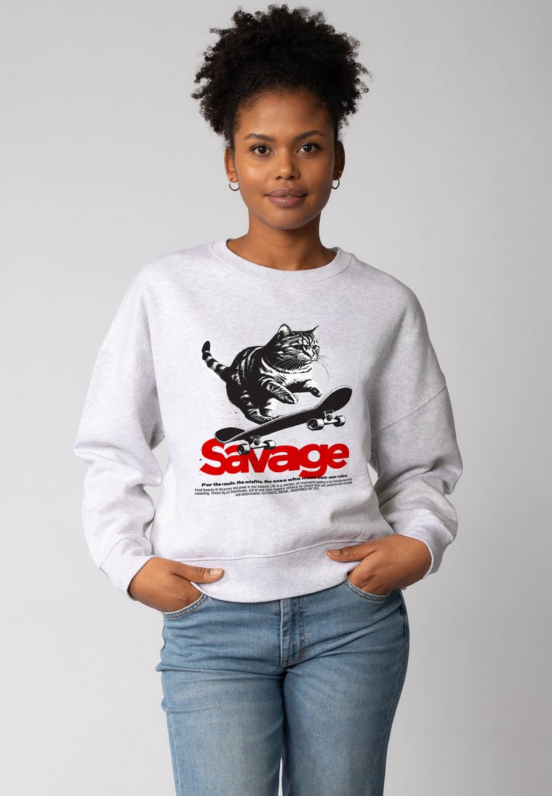 Femme aux cheveux bouclés portant un sweatshirt gris clair avec un chat sur un skateboard, accompagné du texte rouge "Savage", et un jean bleu clair.