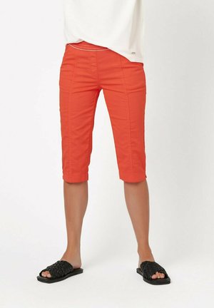 Helle orange Capri-Shorts mit einer körpernahen Passform, sichtbaren Nähten und einem teilweise elastischen Bund. Kombiniert mit schwarzen, geflochtenen Slipper-Sandalen.