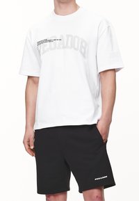 Biała koszulka z bawełny z krótkim rękawem i luźnym krojem, z wyraźnym szarym napisem "PEGADOR" oraz mniejszym czarnym hasłem z przodu.